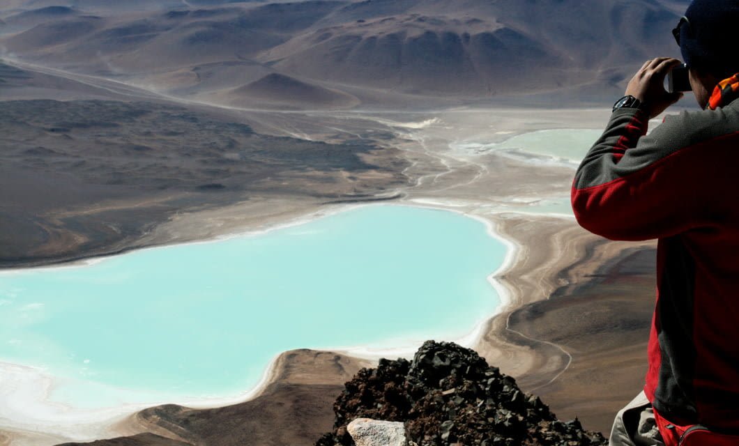 A person in a red and gray jacket stands on a rocky cliff in the Atacama Desert, looking through binoculars at a turquoise lagoon surrounded by arid, mountainous terrain near the Alto Atacama luxury hotel.