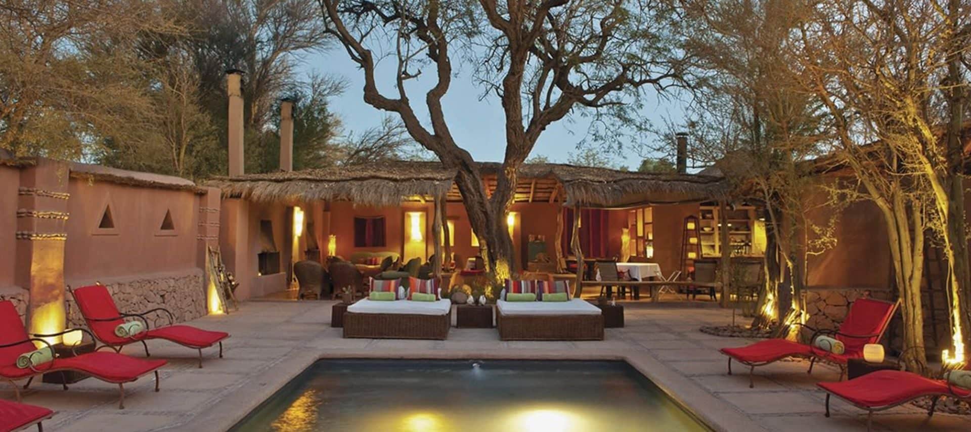 A luxurious outdoor patio with red lounge chairs surrounds a rectangular pool, centered under trees and soft lighting. In the background, an open-walled Chile Lodges building with rustic decor and thatched roof is visible.