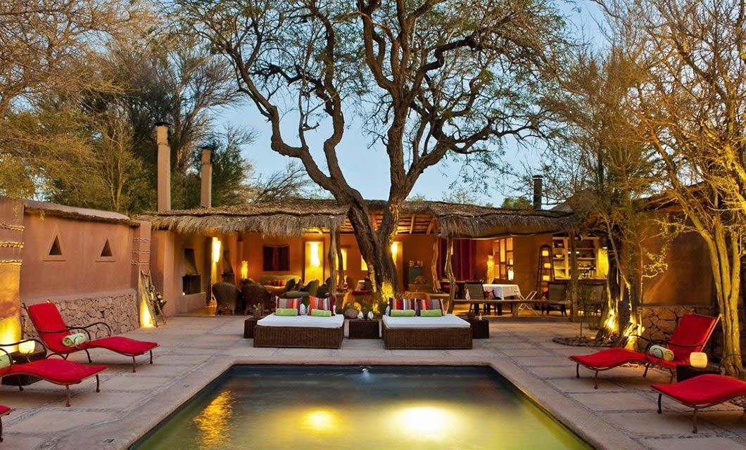 A cozy outdoor patio at dusk features a glowing pool, red lounge chairs, and cushioned daybeds. Nestled in the Atacama Desert, this Awasi Atacama luxury hotel boasts rustic adobe buildings with thatched roofs and open seating areas around a grand tree.