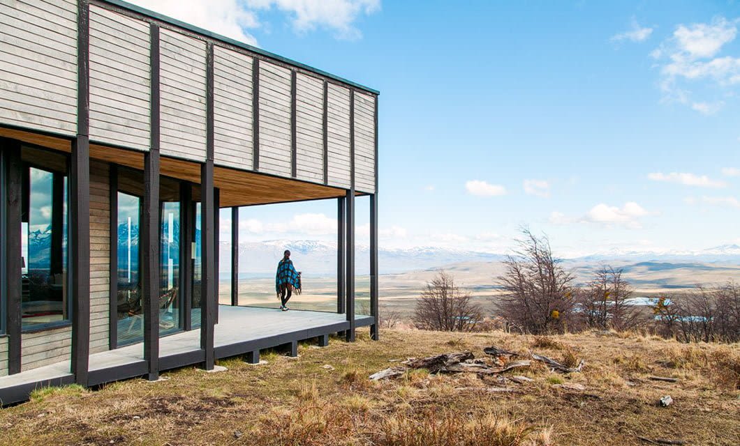 A person wearing a plaid shirt stands on the deck of a modern wooden building at Awasi Patagonia, a luxury hotel in Chile, overlooking a vast landscape with dry grass, scattered trees, and distant mountains under a blue sky with clouds.