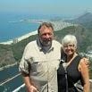 A man and a woman stand smiling together on a scenic overlook, having a wonderful time with the coastline, city, and mountains visible in the background on a sunny day.