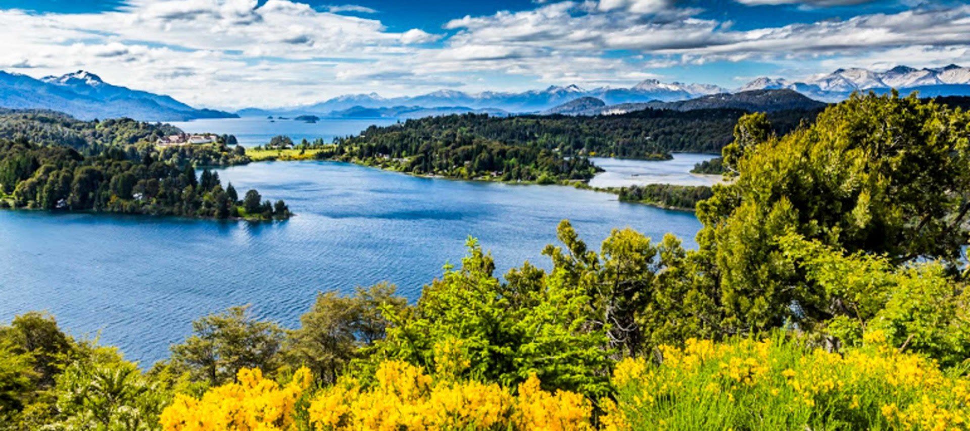 A scenic view of a blue lake in Bariloche, Argentina, surrounded by lush green forests, yellow wildflowers, and distant snow-capped mountains under a partly cloudy sky—perfect for Lake District tours.