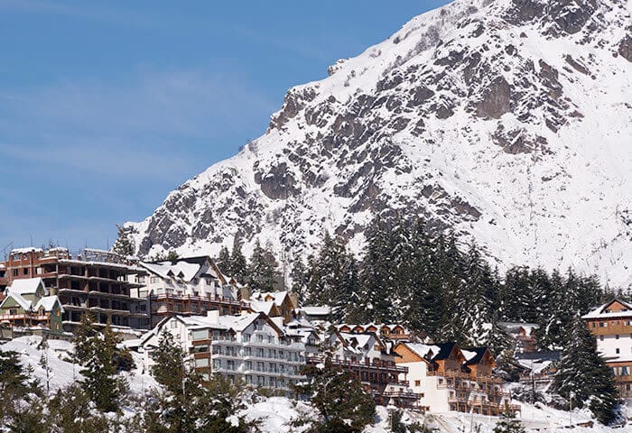 Snow-covered buildings nestled at the base of a large, rugged mountain invite you to discover exciting things to do in this South America travel destination, with snow-capped trees and a clear blue sky in the background.