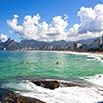 A scenic view of a tropical beach with turquoise water, people swimming, white sand, and distant city buildings backed by lush green mountains under a blue sky with fluffy clouds—perfect for a South America travel adventure.