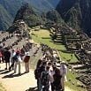 Tourists experience the wonder of Peru as they walk along a path overlooking the ancient stone ruins and terraces of Machu Picchu, set among green mountains under a clear sky.
