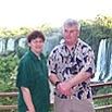 Two people stand in front of a wooden railing with lush greenery and tall waterfalls behind them, both smiling at the camera—ready for their next adventure.