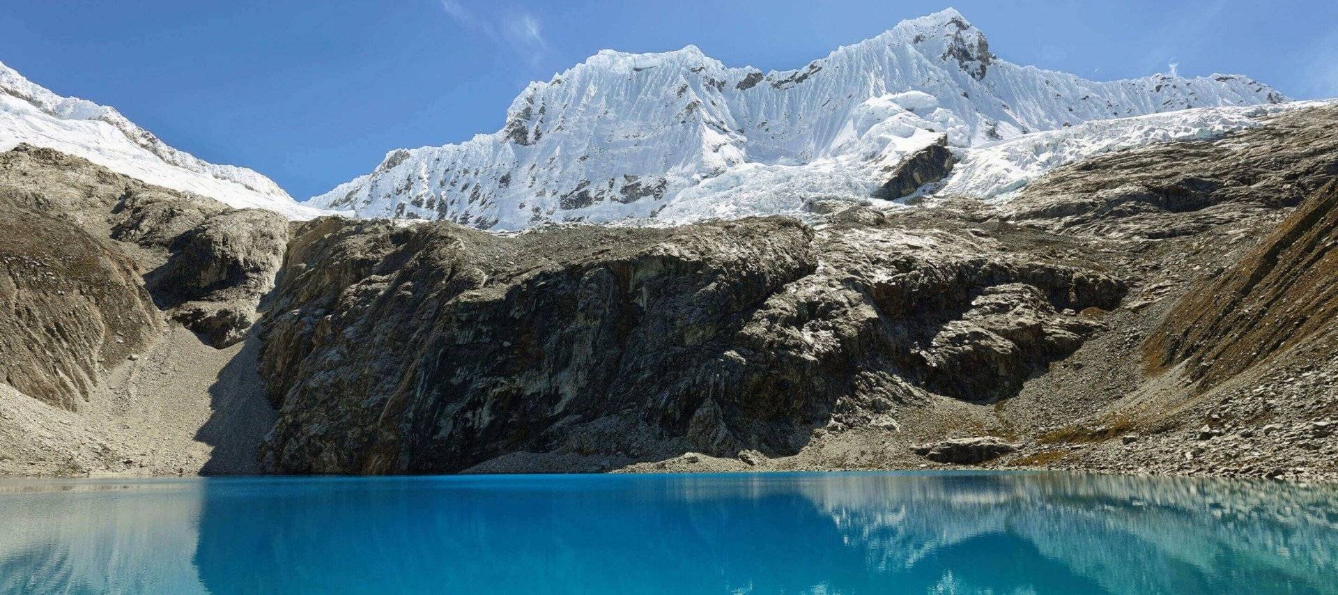 Bright blue lake with clear water in the foreground, rocky shore and steep cliffs in the middle, and tall snow-covered mountains under a clear blue sky—an iconic scene from Huascaran National Park.