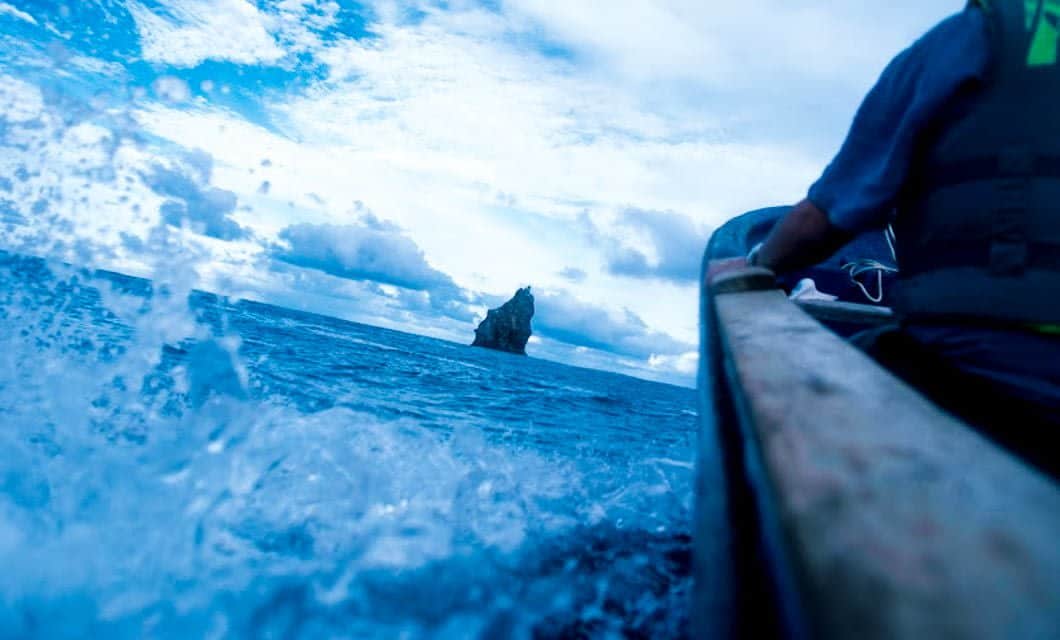 A person in a boat paddles through choppy, blue ocean waters toward a distant, jagged rock formation near Rapa Nui under a partly cloudy sky.