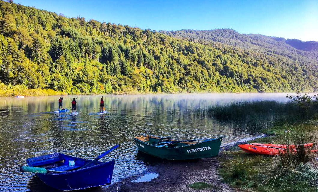 A calm lake surrounded by green forested hills, with three people paddleboarding on the water near Hotel Vira Vira. Several small colorful boats are on the grassy shore in the foreground under a clear blue sky.