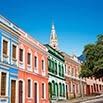 Colorful colonial-style buildings line a street under a bright blue sky, creating an El Dorado of charm, with a church spire in the background and a tree on the right—a truly memorable moment to cherish.