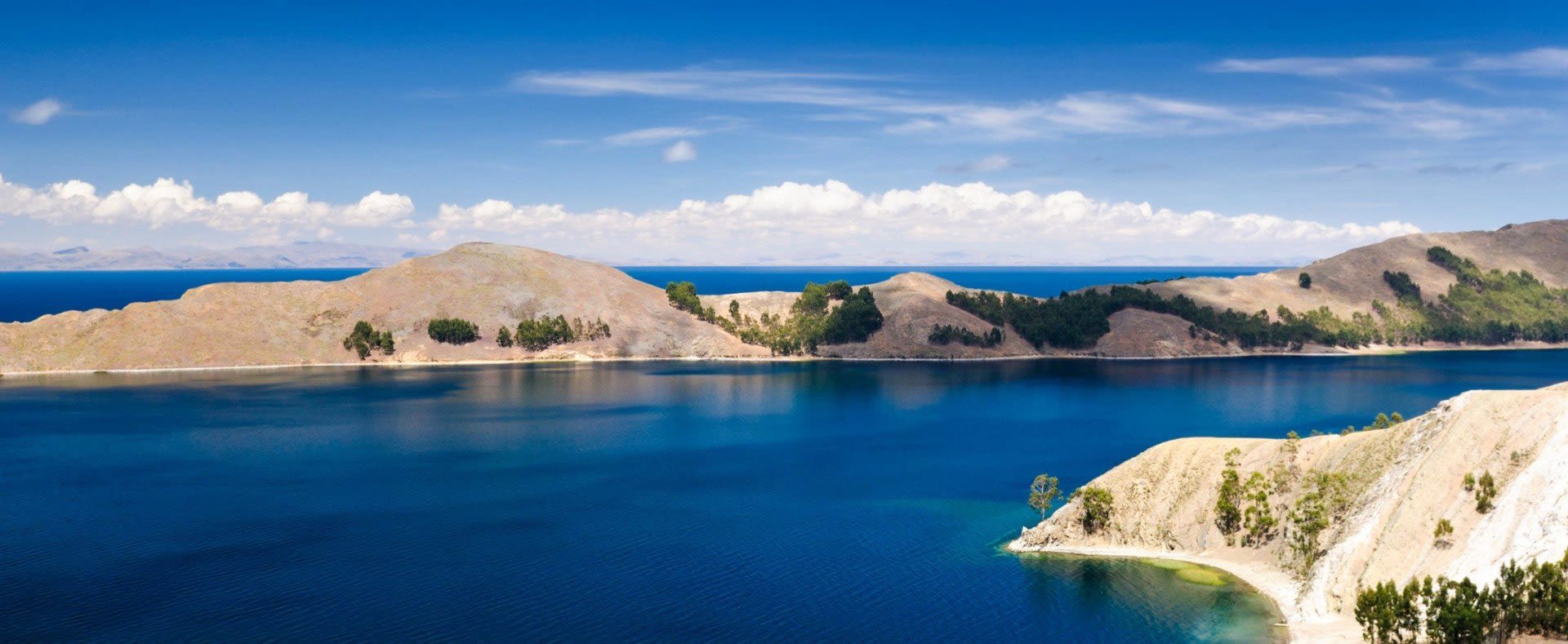 A panoramic view of a deep blue lake in Bolivia, perfect for Bolivia travel, surrounded by gently rolling hills and sparse trees under a clear sky with scattered clouds. The shoreline features sandy and rocky areas.
