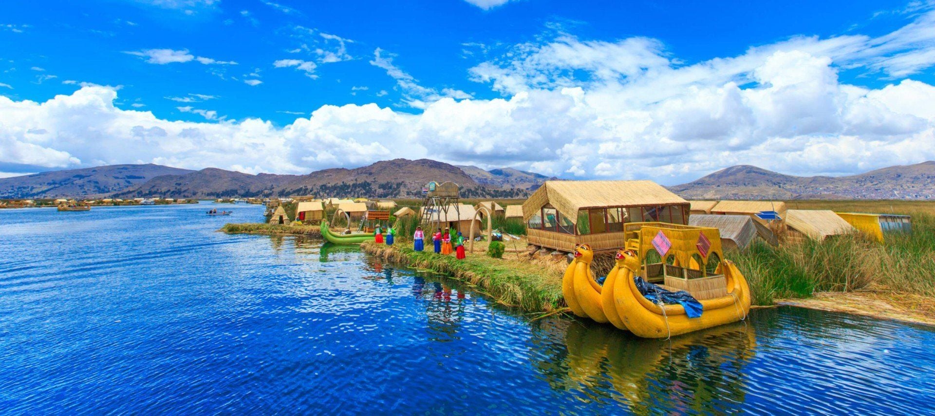 Colorful floating reed houses and boats sit on a bright blue lake with people nearby, backed by mountains and a partly cloudy sky—an iconic scene you can experience on Peru and Bolivia tours.
