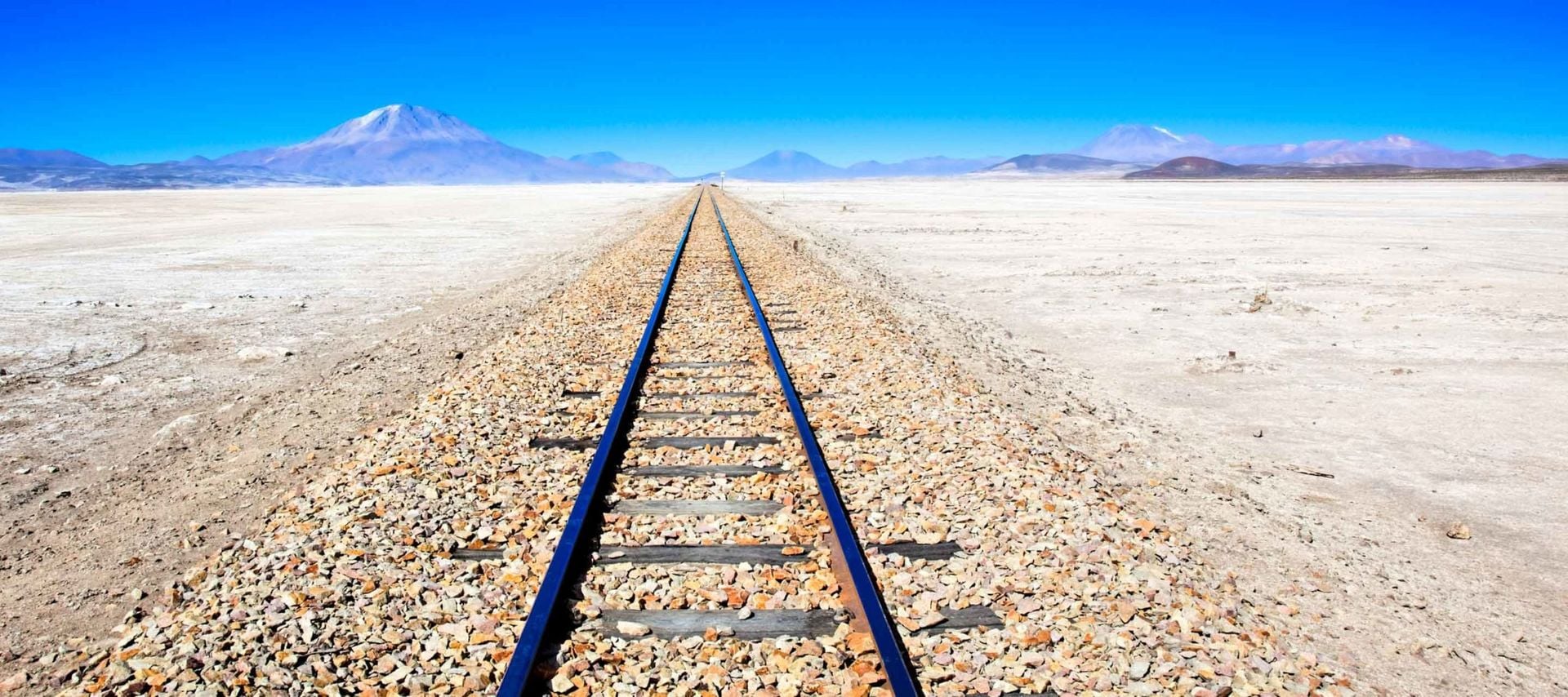 Railway tracks stretch straight into the distance across a barren, rocky desert landscape in Bolivia, creating an iconic scene for train travel Bolivia enthusiasts, with mountains visible on the horizon under a clear blue sky.