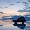 A silhouetted SUV with luggage on top is parked on the Surreal Salt Flats Bolivia, its reflection merging with a dramatic, cloud-filled sunset sky—capturing the mirror effect that defines Bolivia tourism.