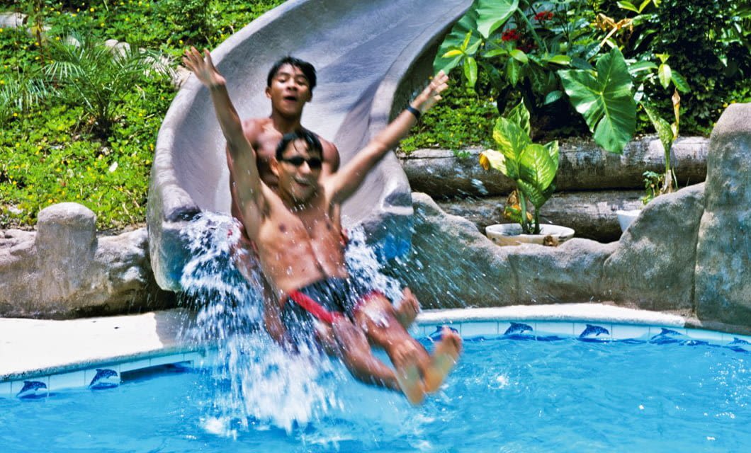 Two people in swimsuits joyfully slide down a water slide into a pool, splashing water at Ceiba Tops Lodge. Lush green plants and trees surround the slide, creating a vibrant tropical atmosphere.