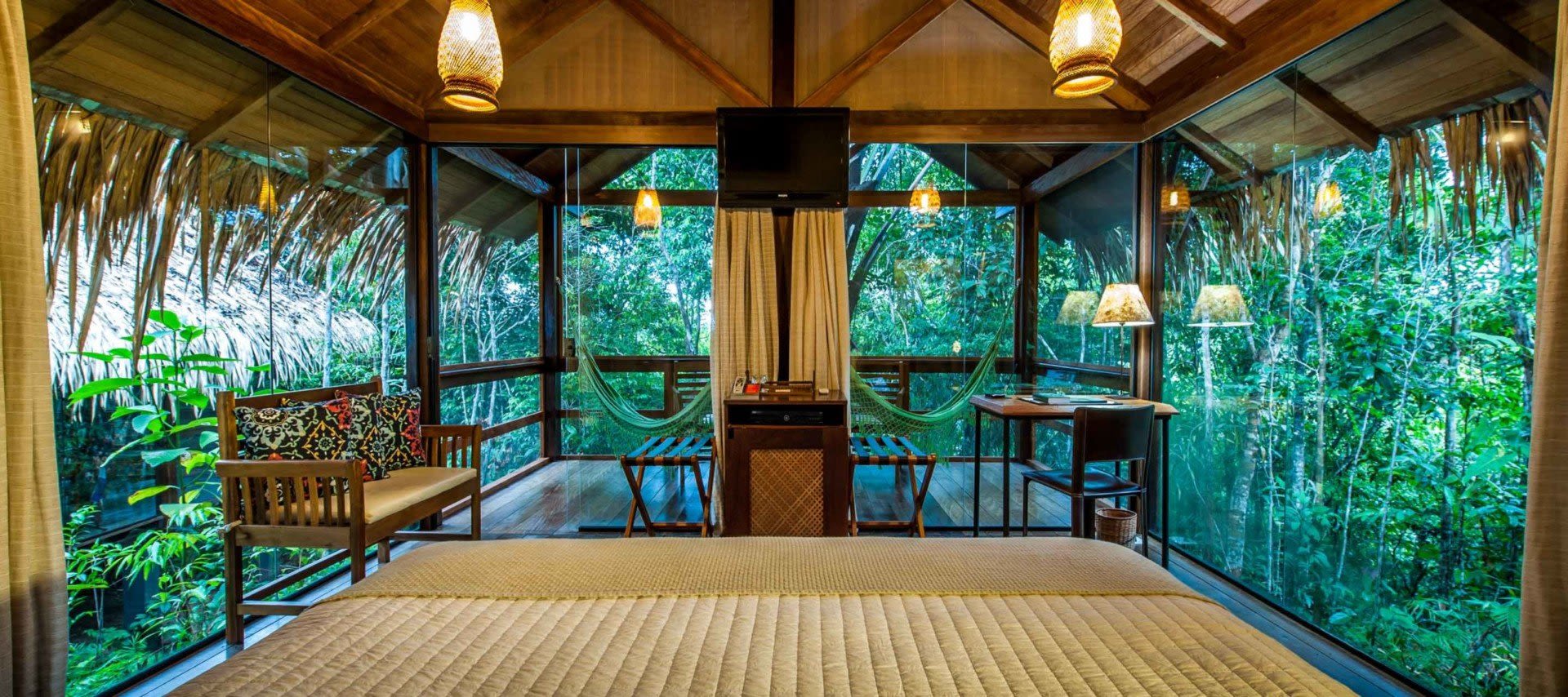 A cozy bedroom in a wooden cabin at an Amazon Lodge, with large glass walls overlooking lush green forest, featuring a bed, bench, hammock, desk, and soft warm lighting from hanging lamps—a perfect Brazil Amazon retreat.