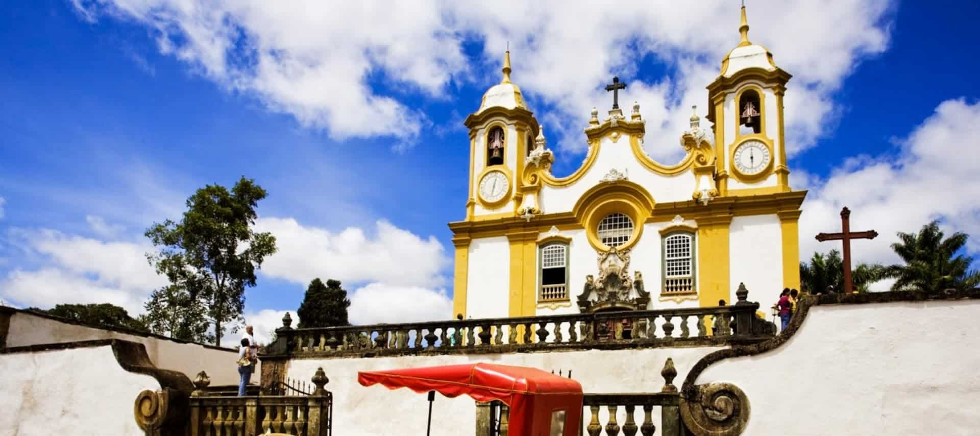 A yellow and white baroque church with twin bell towers stands under a blue sky with clouds, behind a decorative stone fence, with a red tent and trees in the foreground—a highlight for Brazil travel and fans of UNESCO sites.