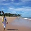 A happy person dressed in white walks along a wide sandy beach with gentle waves, palm trees, and a bright blue sky with scattered clouds—a perfect moment from a memorable trip.