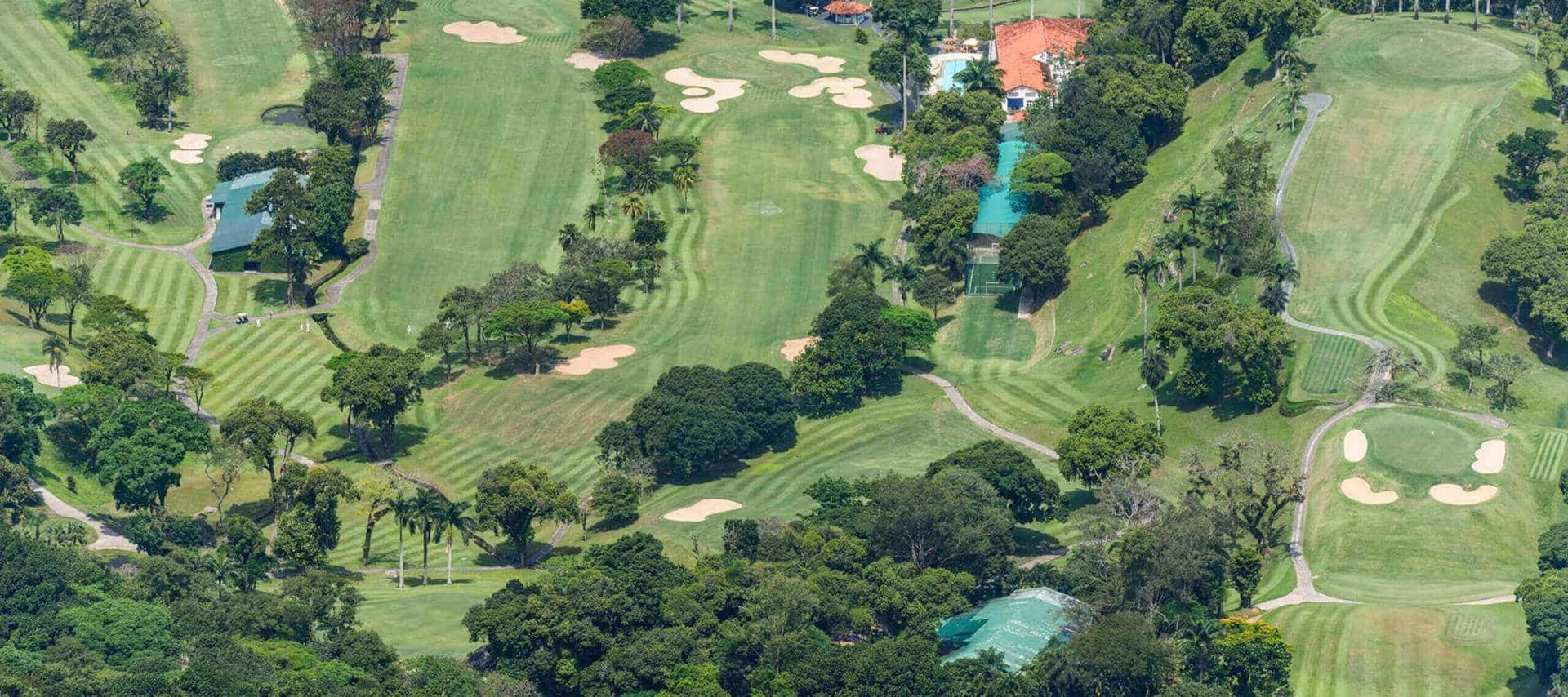 Aerial view of a lush, green golf course ideal for Brazil Golf Tours, featuring winding paths, sand bunkers, ponds, clusters of trees, and nearby buildings with a swimming pool nestled in tropical vegetation.