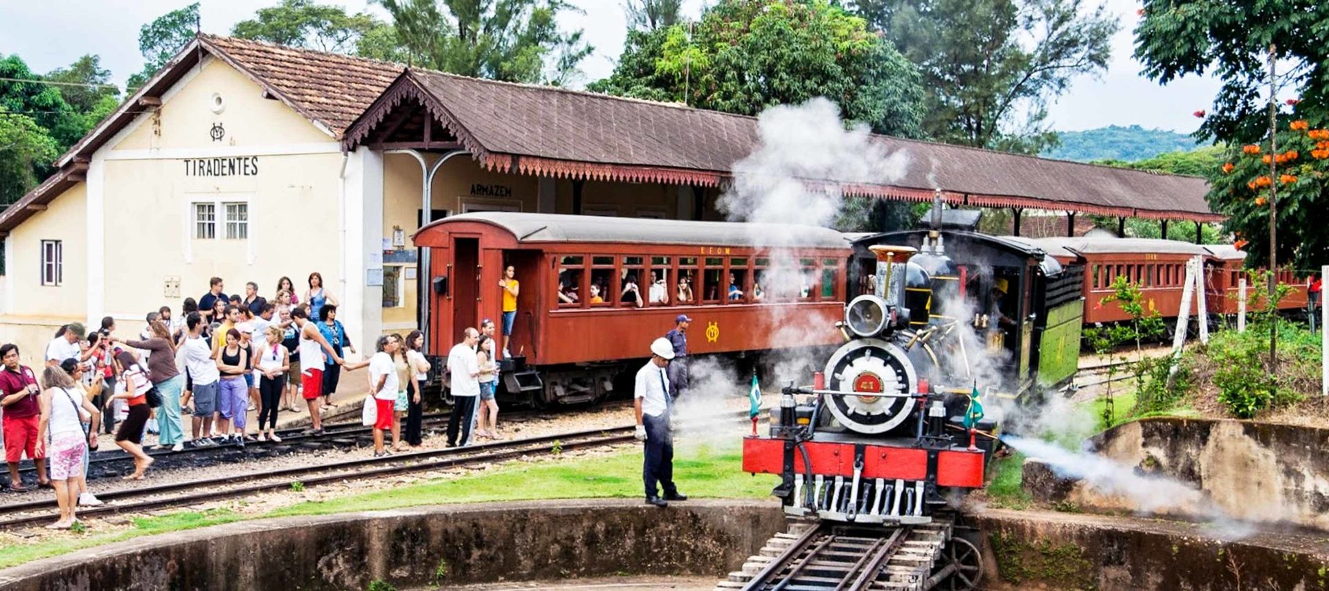 A vintage steam train emits smoke as it arrives at Tiradentes station, part of iconic Brazil Train Tours. A crowd of people, including children, gather to watch and take photos as trees surround the lively rural scene.