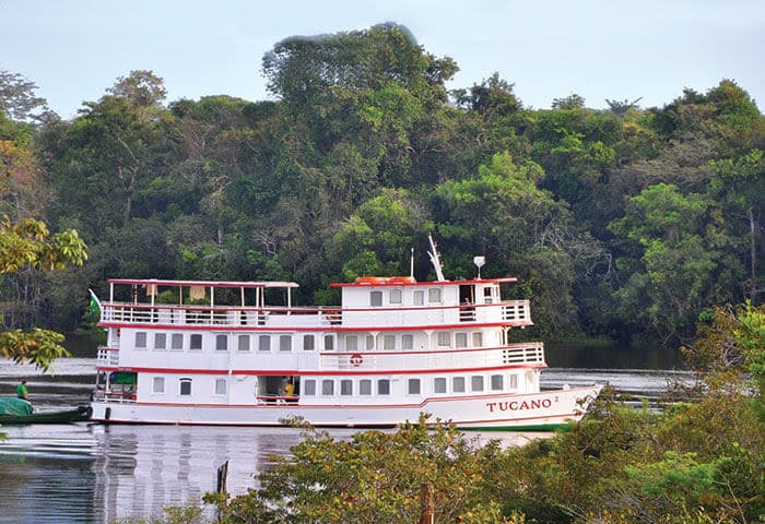 A white riverboat named Tucano with red trim sails on a calm river surrounded by lush green trees and dense forest.