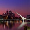 A modern white bridge with a unique angled design spans a calm river at dusk, while city lights sparkle in the water—an unforgettable scene we recommend when exploring with SouthAmerica.travel’s expert travel services.