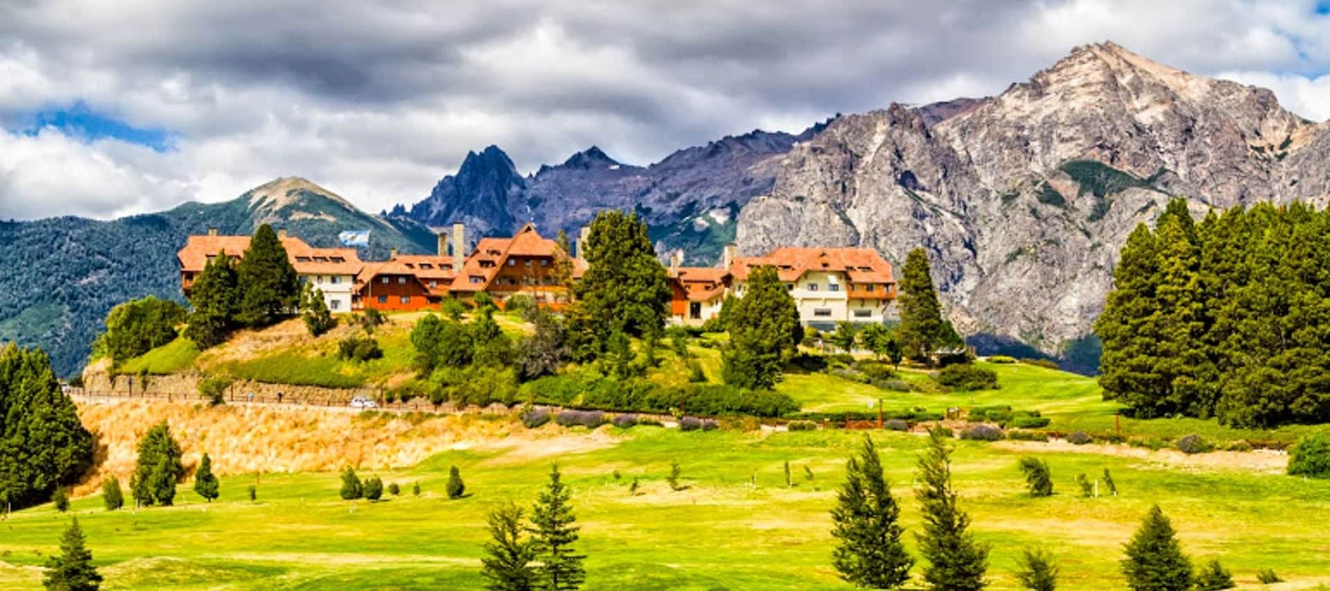 A large building with red-tiled roofs sits on a grassy hill, surrounded by trees, with dramatic mountains and a partly cloudy sky in the background—an idyllic setting often featured in Argentina Golf Tours.