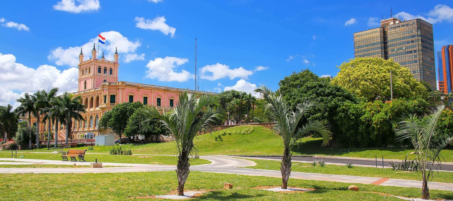 A pink and white historic building with a flag on top stands near palm trees and green lawns under a sunny blue sky, with modern high-rises nearby—an enchanting setting perfect for Paraguay romantic getaways.