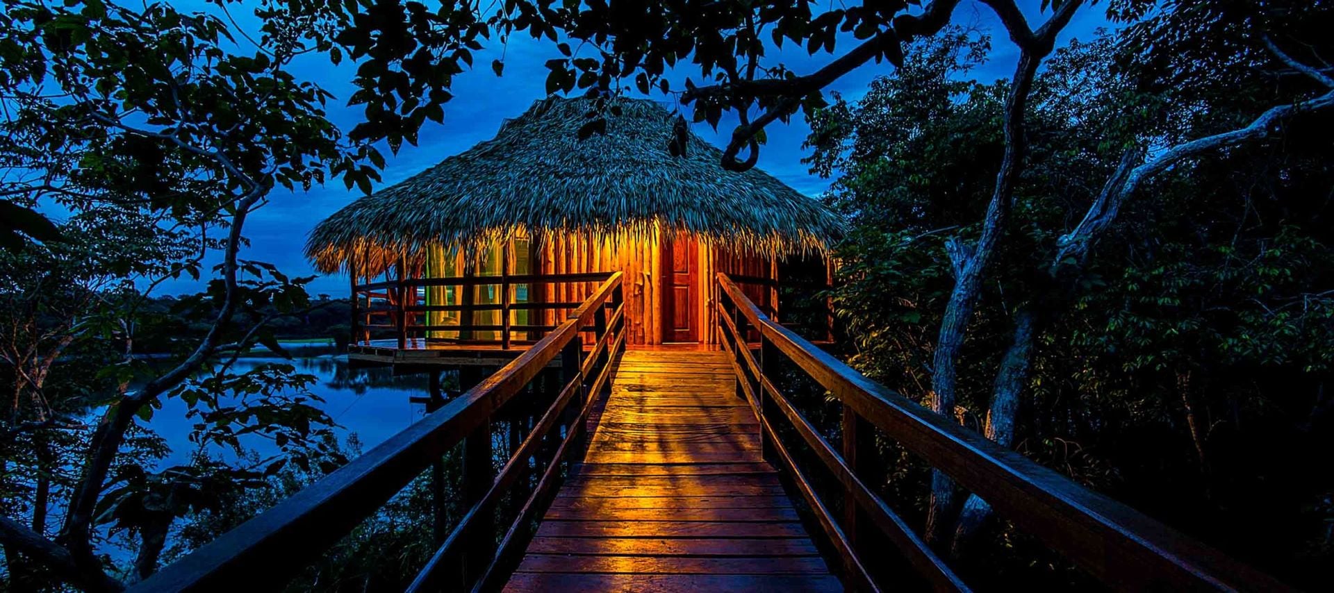 A thatched-roof hut on stilts is warmly lit at night in the style of Amazon Rainforest Lodges, surrounded by lush trees, with a wooden walkway leading toward the glowing entrance over water.