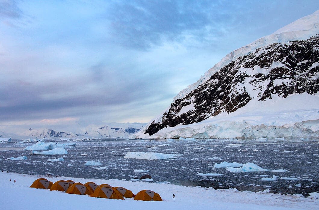 A row of orange tents is set up on snowy ground near the icy sea, with floating icebergs and a towering mountain in the background—the perfect base for unique Antarctica activities and memorable things to do in Antarctica.