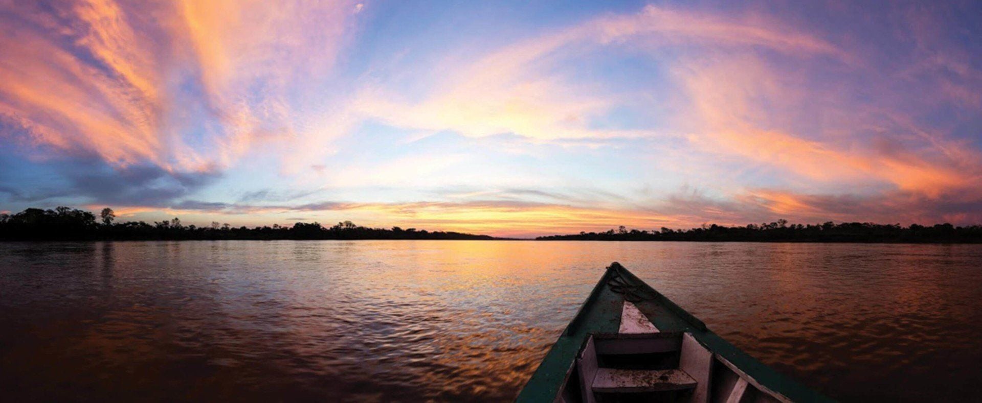 A boat on a calm river at sunset in the Amazon Rainforest, with a vibrant sky of pink, orange, and purple clouds reflecting on the water, and dark silhouettes of trees lining the distant shore—perfect for those arriving on Amazon flights.