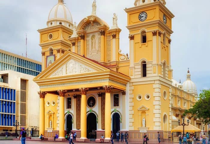 Bright yellow colonial-style cathedral with ornate columns, statues, and two clock towers, surrounded by people and trees; modern buildings rise in the background under a partly cloudy sky—a highlight among Venezuela attractions.