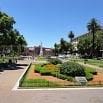 A sunny park in South America with green lawns, manicured bushes, trees, pathways, benches, and a central monument. Buildings line the background under a clear blue sky—a serene spot perfect for travel memories.