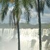 Tall palm trees frame a powerful waterfall cascading over a rocky cliff, with mist rising and a blue sky overhead—an unforgettable sight for an amazing trip or your best vacation travel experience.