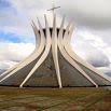 Modern cathedral with curved white columns reaching upward, forming a crown-like structure. A cross sits atop the building against a cloudy sky—an inspiring sight on any wonderful trip. The open, paved surroundings add to its spacious feel.