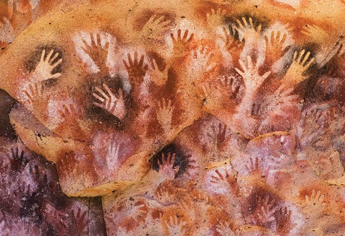 Ancient cave wall covered in numerous hand stencil paintings in various sizes, mostly in reddish-brown and white tones, overlapping and creating a vibrant, textured pattern—one of the unique Argentina attractions for travelers to explore.