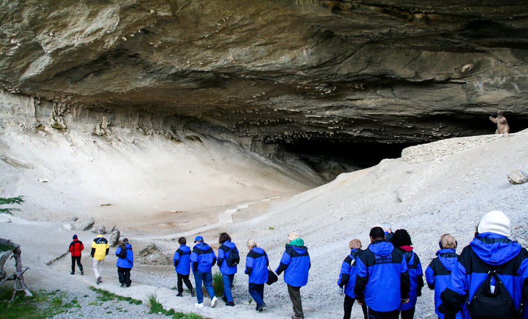 A group of people wearing blue jackets walk towards the entrance of a large cave with a rocky ceiling near Lago Grey Lodge; a single animal stands on a ledge by the cave opening.