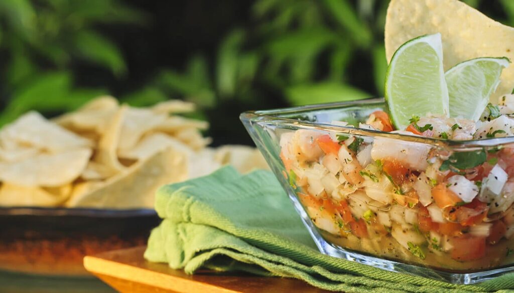 A glass bowl of fresh ceviche—a must among foods to try in Lima Peru—with diced tomatoes, onions, cilantro, and shrimp, garnished with lime slices and a tortilla chip. A pile of tortilla chips is blurred in the background.