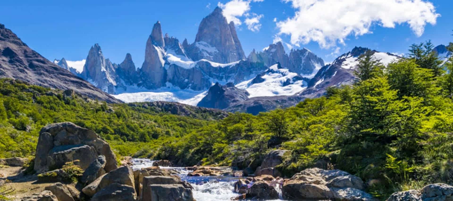 Snow-capped jagged mountain peaks rise above lush green forest and a rocky stream under a bright blue sky with scattered white clouds—perfect for scenic tours in Argentina’s breathtaking UNESCO sites.