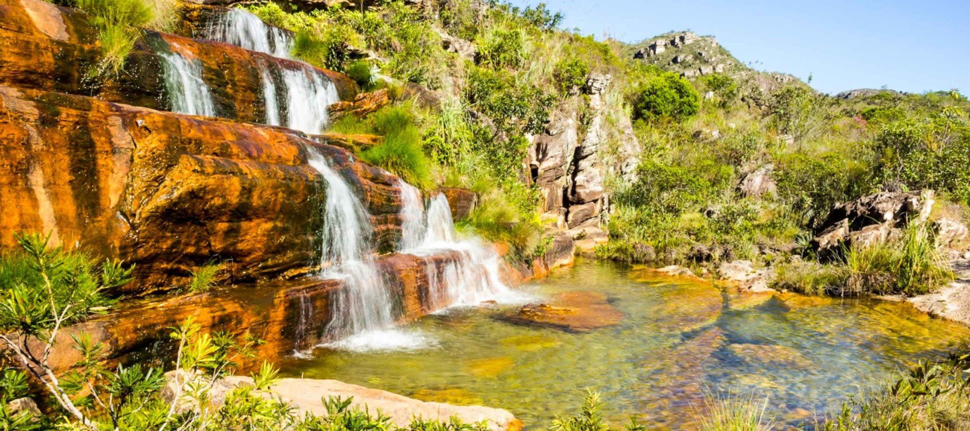 Small waterfalls flow over reddish rocks into a clear, shallow pool surrounded by green plants and shrubs, with hills and a blue sky in the background—a picturesque scene often explored on Chapada Diamantina Tours.