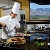 A chef in a white uniform and tall hat slices meat on a wooden board in a modern kitchen with mountain views. Fresh ingredients and utensils line the counter, reflecting the highly recommended spot's great service and culinary excellence.