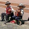 During an Argentina trip in April-May, two young children wearing hats and formal clothes sit on small chairs outdoors, playing red accordions. They appear to be performing on a paved street with a brick wall in the background.