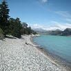 A rocky shoreline with smooth grey stones leads to a turquoise lake, perfect for your Argentina trip. Tall evergreen trees line the shore on the left, and distant mountains rise under a partly cloudy blue sky.