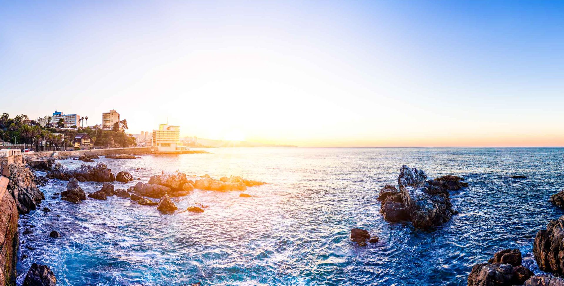 A panoramic view of Chile’s rocky coastline at sunrise or sunset, with waves crashing against the rocks and buildings lining the shoreline under a clear blue sky—an ideal scene for any travel guide.