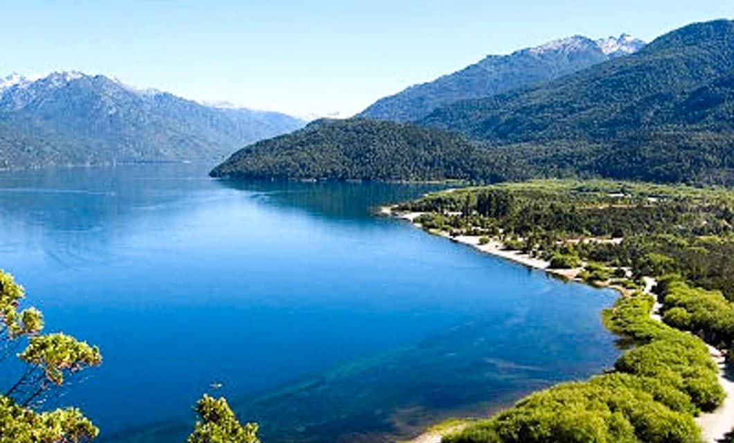 A large, calm blue lake surrounded by lush green forests and mountains under a clear blue sky, with the shoreline curving gently into the distance—an idyllic scene near Mitico Puelo Lodge.