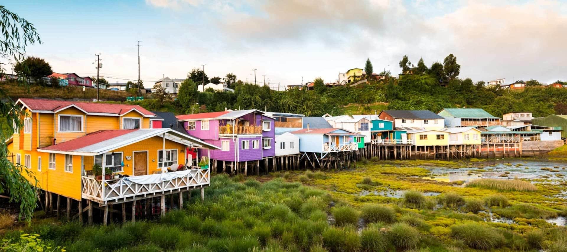 Colorful wooden houses on stilts stand above grassy wetlands on Chiloe Island, with a hillside and more houses in the background under a cloudy sky.