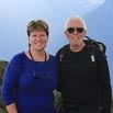 Two people are standing outdoors, smiling at the camera during a trip of a lifetime. The woman on the left is in a blue top, while the man on the right sports sunglasses, a black shirt, and a backpack. Majestic mountains rise in the background.