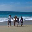 A family of four walks hand in hand along a sandy beach, with blue ocean waves and a clear sky in the background—an experience you'd recommend to a friend.