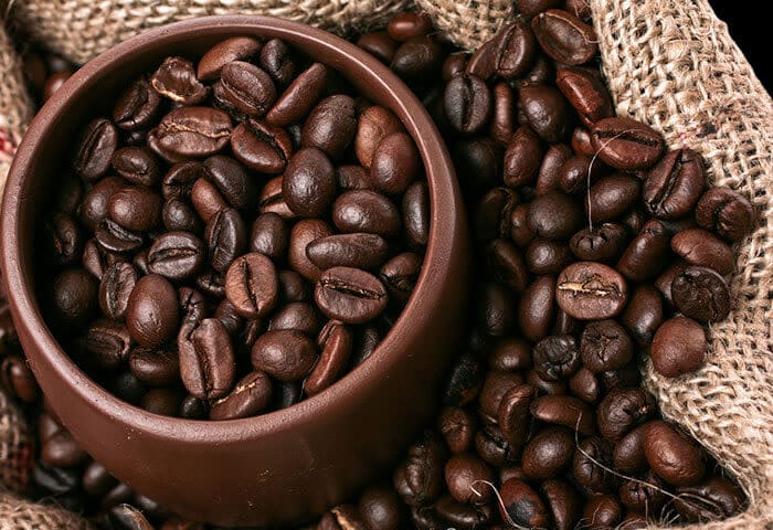 A brown ceramic bowl filled with roasted coffee beans sits on a burlap sack, with more beans scattered around it—a perfect glimpse into South America travel and one of the region’s must-try activities for coffee lovers.