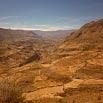 A wide, arid canyon landscape with rugged, rocky hills and a clear blue sky offers a smooth experience for explorers. The terrain is mostly brown and dry, stretching towards distant mountains on the horizon.